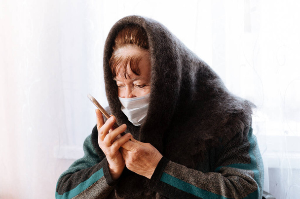 Una nonna anziana con una maschera medica chirurgica e un fazzoletto con un telefono in mano, quarantena, auto-isolamento, coronavirus
 - Foto, immagini