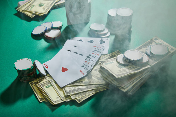 High angle view of playing cards, dollar banknotes and casino tokens with smoke around on green - 写真・画像