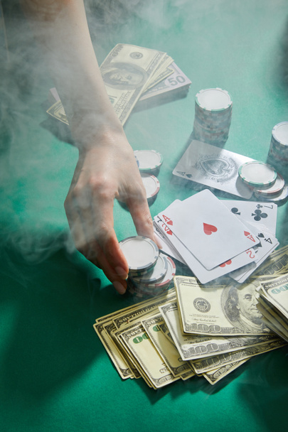 Cropped view of female hand with casino tokens, playing cards and money with smoke around on green background - Foto, immagini