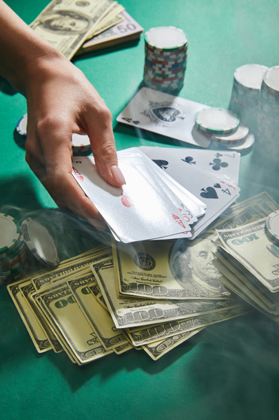 Cropped view of woman holding playing cards near dollar banknotes and casino tokens with smoke around on green  - 写真・画像