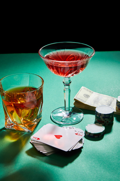 Glasses of cognac and cocktail near dollar banknotes, casino tokens and playing cards on green surface isolated on black - Fotó, kép