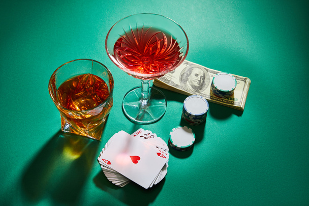 High angle view of glasses of cognac and cocktail near dollar banknotes, casino tokens and playing cards on green - Foto, Bild