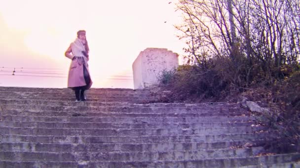 A young girl in a pink coat goes down the stairs in the park on autumn sunny evening. Stock footage. Retro landscape with a beautiful woman walking in a city park. - Footage, Video