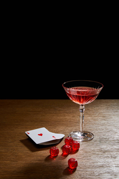 Glass of cocktail near deck of cards and dice on wooden surface isolated on black - Foto, Imagen