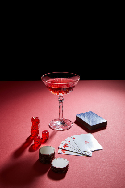 Glass of cocktail near playing cards, casino chips and dice on red surface isolated on black - Foto, immagini