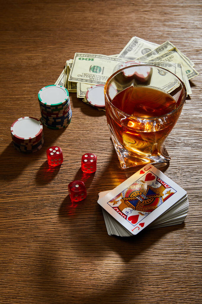 High angle view of glass of cognac near dollar banknotes, playing cards, casino chips and dice on wooden background - Fotó, kép