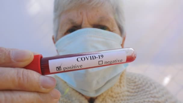 Korumalı maskeli yaşlı kadın kan testini tutuyor ve koronavirüs pozitif sonuç veriyor. Hasta nine üzgün duygular gösteren kameraya bakıyor. Salgın COVID-19 'dan sağlık ve güvenlik hayatı kavramı - Video, Çekim