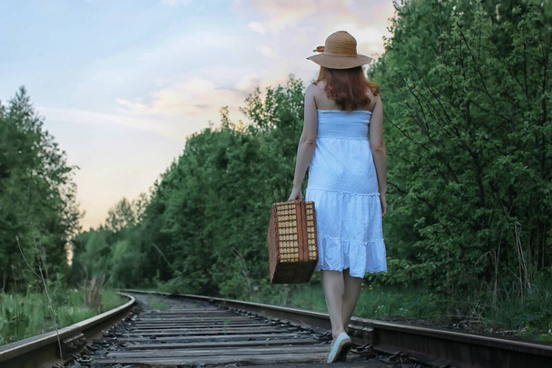 meisje in een witte sundress en een rieten koffer lopen op rail - Foto, afbeelding