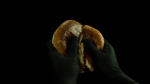 Close up de mão em luvas de cozinha pretas quebrando pão em dois pedaços isolados em fundo preto. Imagens de stock. Conceito de comida e culinária, pão de hambúrguer fresco sendo dividido
. - Foto, Imagem