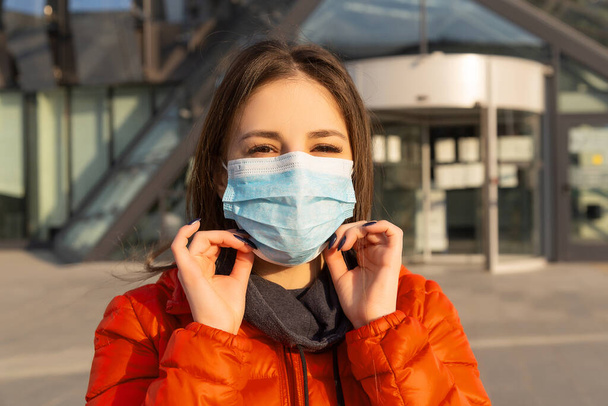 A woman wears a protective mask against allergies, viruses, air pollution. Climate change concept. Portrait of a young woman in a medical mask from a coronavirus in a red jacket on the street - Foto, Imagem