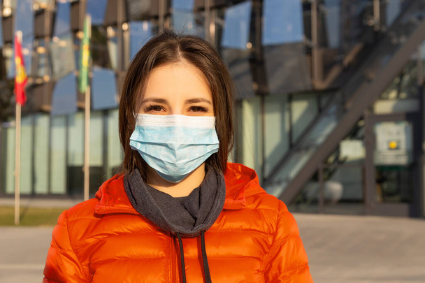 Una mujer lleva una máscara protectora contra alergias, virus, contaminación del aire. Concepto de cambio climático. Retrato de una joven con una máscara médica de un coronavirus en una chaqueta roja en la calle
 - Foto, Imagen