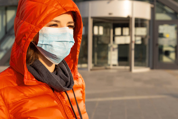 女性はアレルギー、ウイルス、大気汚染に対する保護マスクを着用しています。気候変動の概念。通りの赤いジャケットのコロナウイルスからの医療用マスクの若い女性の肖像画 - 写真・画像