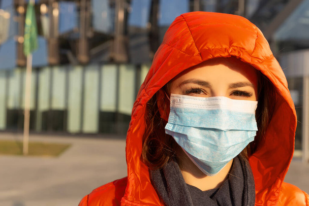 Uma mulher usa uma máscara protetora contra alergias, vírus, poluição do ar. Conceito de mudança climática. Retrato de uma jovem mulher em uma máscara médica de um coronavírus em uma jaqueta vermelha na rua
 - Foto, Imagem
