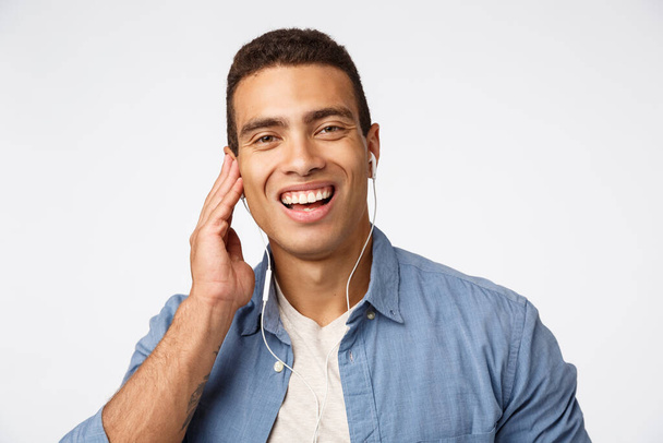 Close-up cheerful, happy laughing young handsome man in casual outfit, put headphones, touch earphone and smiling as joyfully talking with somone on way home, listen music, favorite songs or podcast - Photo, Image