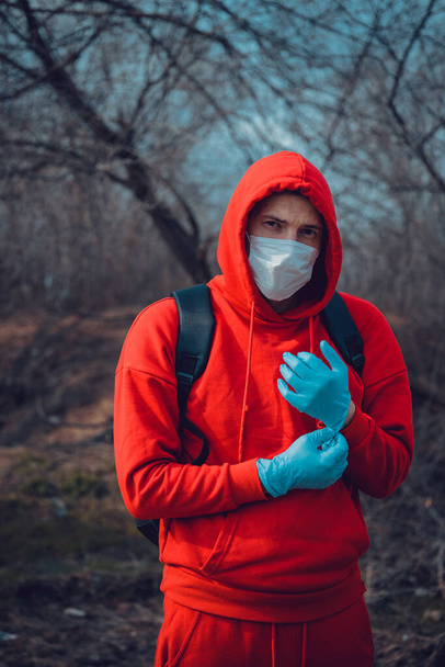 Young man in medical mask putting on gloves in countryside. Adult male protecting yourself from diseases on walk. Concept of threat of coronavirus epidemic infection. - Фото, изображение