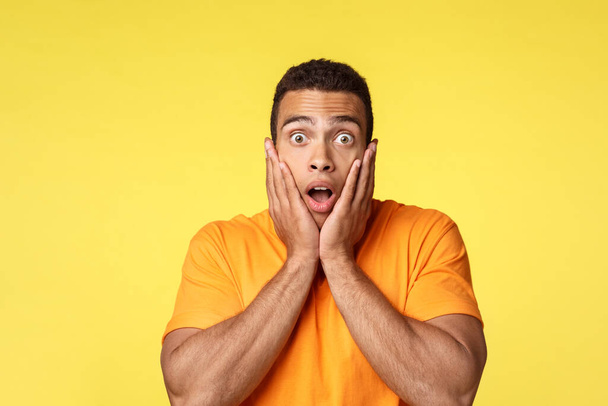 Surprised and speechless young shocked handsome man in t-shirt, gasping amazed, drop jaw and stare camera popped eyes, grab face from amazement, standing stupor from realisation, yellow background - Photo, Image