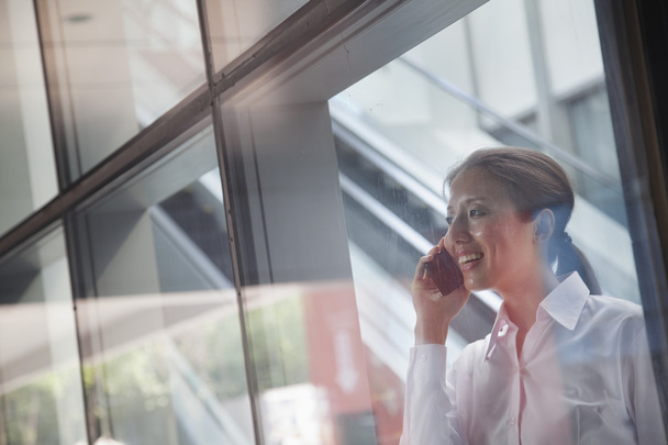 Joven mujer de negocios dentro del edificio hablando por teléfono y mirando a través del vidrio
 - Foto, Imagen