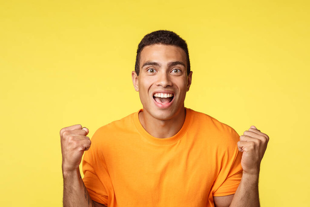 Achievement, celebration and win concept. Handsome relieved cheerful young man clench hands, fist pump and smiling become winner or champion, receive prize, achieve success, yellow background - Фото, зображення