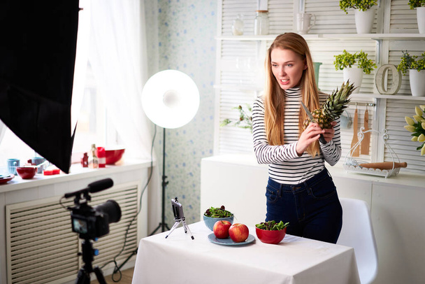Food blogger cooking fresh vegan salad of fruits in kitchen studio, filming tutorial on camera for video channel. Female influencer holds apple, pineapple and talks about healthy eating. Fructorianism - Foto, Imagen