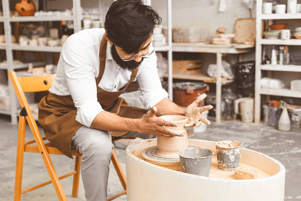 A young male potter is engaged in craft in his workshop on a potters wheel and makes a clay product - Fotografie, Obrázek