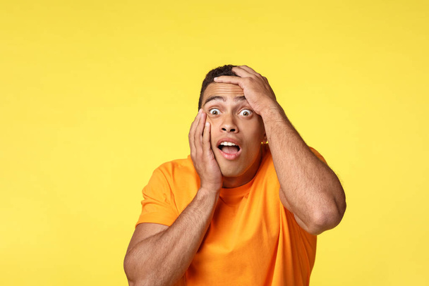 Scared, stunned and speechless frightened handsome man, grab head gasping, open mouth and stare away terrified, feeling fear and discomfort, trembling from horror, stand yellow background - Photo, Image