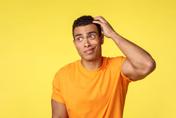 Unsure, clumsy young slightly dumb guy in casual t-shirt, scratching head clueless smirk and look away troubled answer, cant tell feeling confused and unsure, have no clue, stand yellow background - Photo, Image