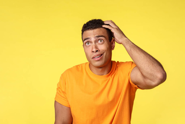 Indecisive pensive handsome silly guy in t-shirt, scratch head and look up confused, smirk as trying figure out something, cant remember or dont have ideas, standing questioned yellow background - Photo, Image