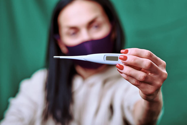 a brunette girl wearing a purple medical mask shows a white electronic thermometer into the camera. green background. covid-19. protect the health. hygiene and quarantine. check of temperature - 写真・画像
