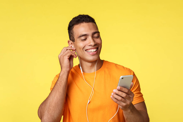 Joyeux jeune homme masculin souriant en t-shirt orange, tenant smartphone, bavardant en écoutant de la musique ou en regardant la vidéo dans un casque, a reçu un message vocal et en riant dessus, fond jaune
 - Photo, image