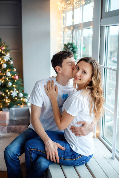 Κάθετη προσωπογραφία του άνδρα και της γυναίκας στο σπίτι την παραμονή των Χριστουγέννων. Χαρούμενη οικογενειακή φίλη και το αγόρι κοντά στο χριστουγεννιάτικο δέντρο κάθεται στο περβάζι παράθυρο το χειμώνα. Άντρας φιλάει γυναίκα στο μάγουλο - Φωτογραφία, εικόνα