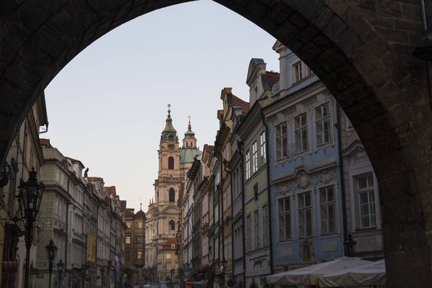 As seen from an archway in a Prague street, the historic building facades are ornate and cream colored and the roofs have pointed greenish domes - Foto, afbeelding