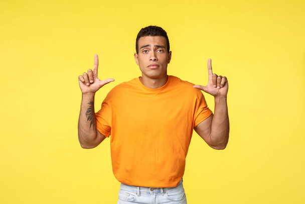 Грустный мрачный молодой кавказский мужчина в оранжевой футболке хмурясь расстроен, указывая на обиженные или огорченные, теряя конкуренцию, чувствуя печаль и депрессию, стоя разочарованный на желтом фоне
 - Фото, изображение