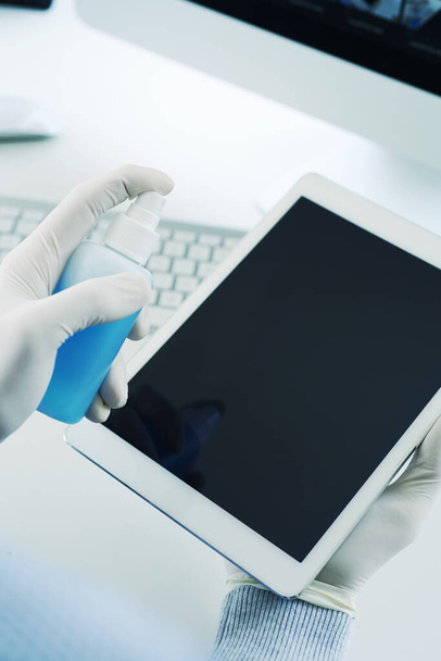 close-up de um homem, usando luvas de látex sentado em uma mesa, desinfetando seu tablet digital por pulverização de um desinfetante azul de uma garrafa
 - Foto, Imagem