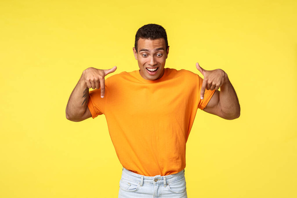 Vzrušený pohledný hispánský chlapík, oblečený do oranžového trička, bílých kalhot, s nadšením a dojmem pozvedne obočí, radostně se usměje, ukazuje a dívá se dolů překvapeně - Fotografie, Obrázek