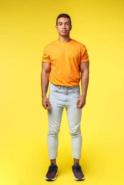 Full-length vertical shot masculine hipster guy, hispanic ethnicity, standing casually yellow background, wear stylish orange t-shirt, white pants, look camera with no expression, slightly smiling - Photo, Image