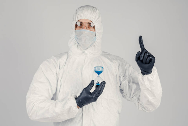 Ένας άντρας με λευκή προστατευτική στολή, μια μάσκα σε λευκό φόντο, με ένα ρολόι στα χέρια του. Μια επιδημία πανδημίας είναι ένας νέος ιός ταχείας εξάπλωσης, μια ιατρική έννοια - Φωτογραφία, εικόνα