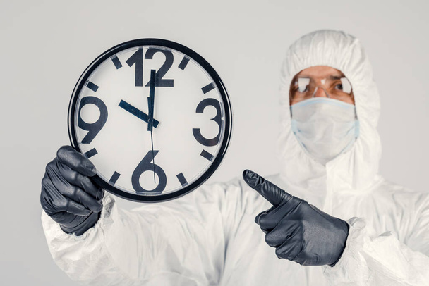 Ένας άντρας με λευκή προστατευτική στολή, μια μάσκα σε λευκό φόντο, με ένα ρολόι στα χέρια του. Μια επιδημία πανδημίας είναι ένας νέος ιός ταχείας εξάπλωσης, μια ιατρική έννοια - Φωτογραφία, εικόνα