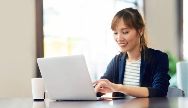 Szczęśliwa młoda bizneswoman korzystająca z laptopa robi zakupy online w kawiarni. Uśmiechnięta piękna Azjatka siedząca i pracująca w swoim miejscu pracy. Styl życia, Technologia, Internet, Komunikacja bezprzewodowa. - Zdjęcie, obraz