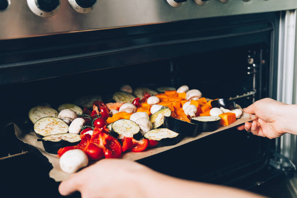 Dłonie kobiety umieszczając surowe warzywa na tacy do piekarnika do pieczenia. Zdrowa kolacja Detox z bakłażana, czerwona papryka, pomidory wiśniowe, dynia - Zdjęcie, obraz