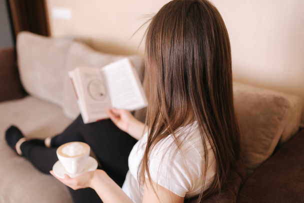 Joven mujer atractiva leer el libro en casa. Hora de la cuarentena. Mujer beber cappuchino y llenar el cuaderno
 - Foto, imagen