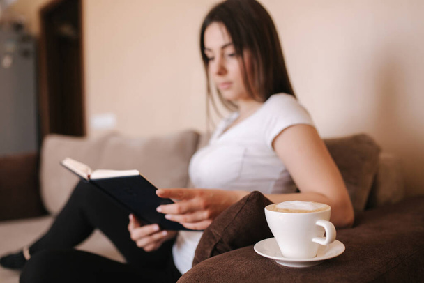 Młoda, atrakcyjna kobieta czyta książkę w domu. Czas kwarantanny. Kobieta pije cappuchino i wypełnia zeszyt - Zdjęcie, obraz