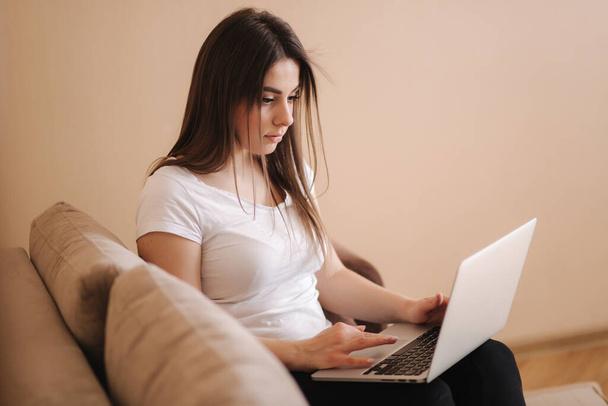 Una joven trabaja en casa en cuarentena. La mujer usa el portátil en el sofá. Mujer estudio a distancia aprendizaje
 - Foto, Imagen