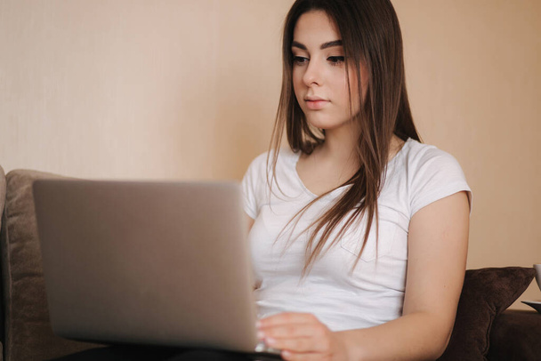 Una joven trabaja en casa en cuarentena. La mujer usa el portátil en el sofá. Mujer estudio a distancia aprendizaje
 - Foto, Imagen