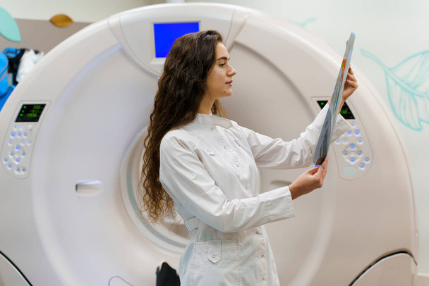 Η νεαρή φοιτήτρια ιατρικής με ρόμπα κοιτάζει την ακτινογραφία του εγκεφάλου μετά από αξονική τομογραφία. CT διαγνωστική στην ιατρική κλινική - Φωτογραφία, εικόνα