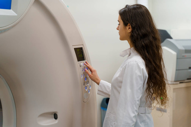 Médico faz tomografia computadorizada paciente fot. Menina jovem de casaco branco olha na câmera e pressione o botão no CT
 - Foto, Imagem