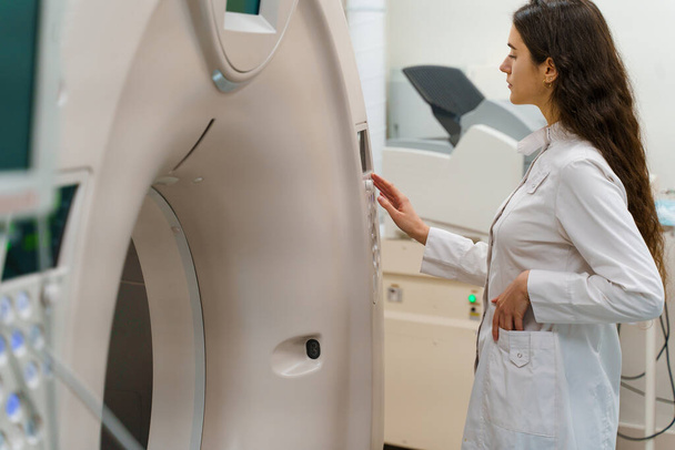 Lékař dělá počítačovou tomografii pacienta. Mladá dívka v bílém plášti hledá v CT a stiskněte tlačítko pro zahájení postupu výzkumu diagnostika - Fotografie, Obrázek