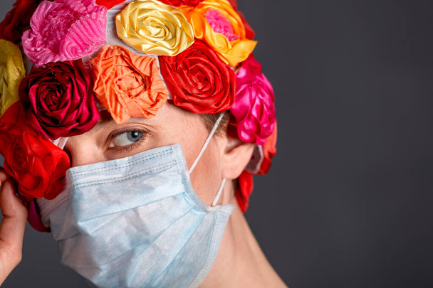 Frau mit medizinischer Maske mit künstlichem Blumenhut vor grauem Hintergrund. Kopierraum - Foto, Bild