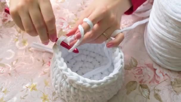 Young woman while crocheting, close up. Leisure activity. White basket made of white T-shirt yarn, with red crochet needle. Pale pink nails. Elegant tablecloth with floral pattern. - Footage, Video