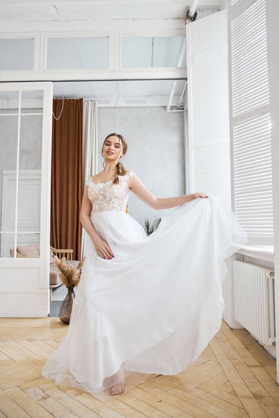 Bride is a woman in a light summer wedding dress standing at the window. Brown-haired girl with perfect hair and beautiful makeup - Foto, imagen