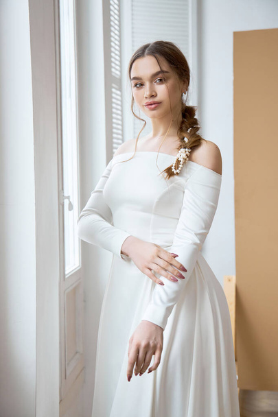 Beautiful slender blonde in the evening sun in a long white dress near a large window. Perfect hair and makeup for the bride, a new collection of wedding dresses - Foto, afbeelding